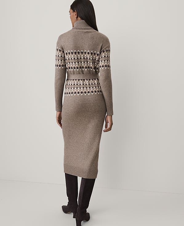 A woman stands with her back to the camera wearing a brown patterned knit dress and dark boots against a plain white background.