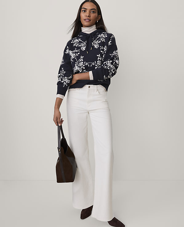 A woman wearing a floral navy hoodie and white flared pants stands against a plain light gray background.