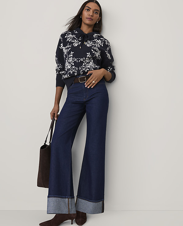 A woman wearing a floral navy sweatshirt and wide-leg blue jeans stands against a plain, light-colored background.