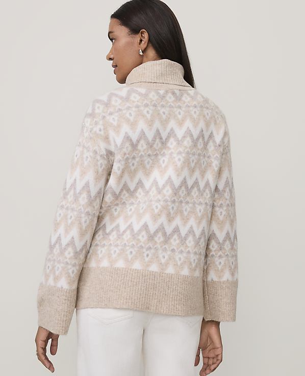 A woman wearing a beige and white patterned turtleneck sweater and white pants stands against a plain light gray background.