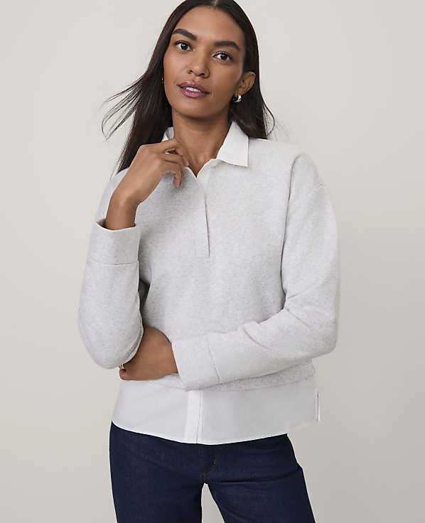 A woman wearing a light grey sweater over a white collared shirt and dark blue jeans stands against a plain, light-colored background.