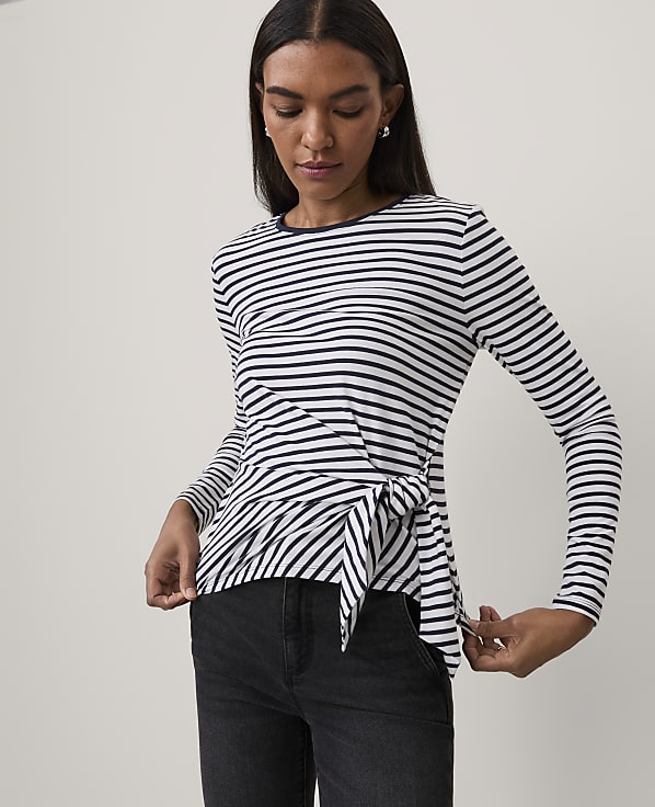 A woman wearing a navy and white striped long-sleeve top with a tie-front detail and black jeans stands against a plain white background.