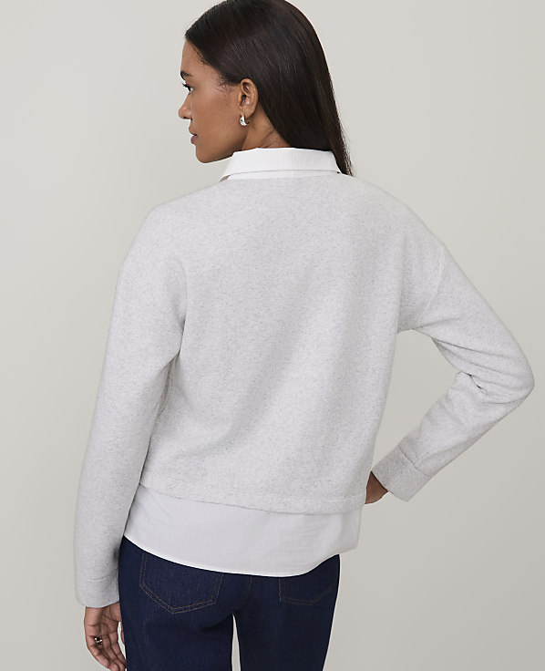A woman wearing a light grey sweater over a white shirt and dark blue jeans stands against a plain, neutral background.
