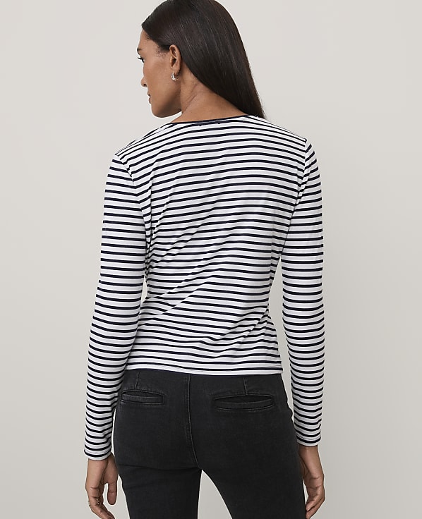 A woman wearing a black and white striped long-sleeve shirt and black jeans stands against a plain light gray background.