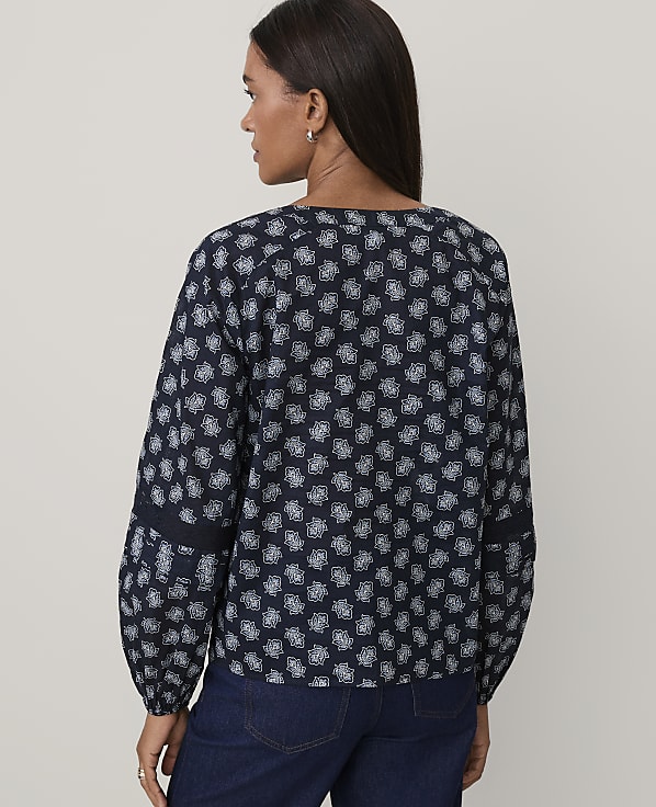 A woman wearing a navy blue patterned blouse and dark blue jeans stands against a plain, light-colored background.