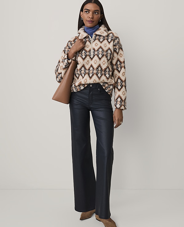 A woman wearing a patterned fuzzy pullover, dark wide-leg trousers, and brown boots stands against a plain light gray background.