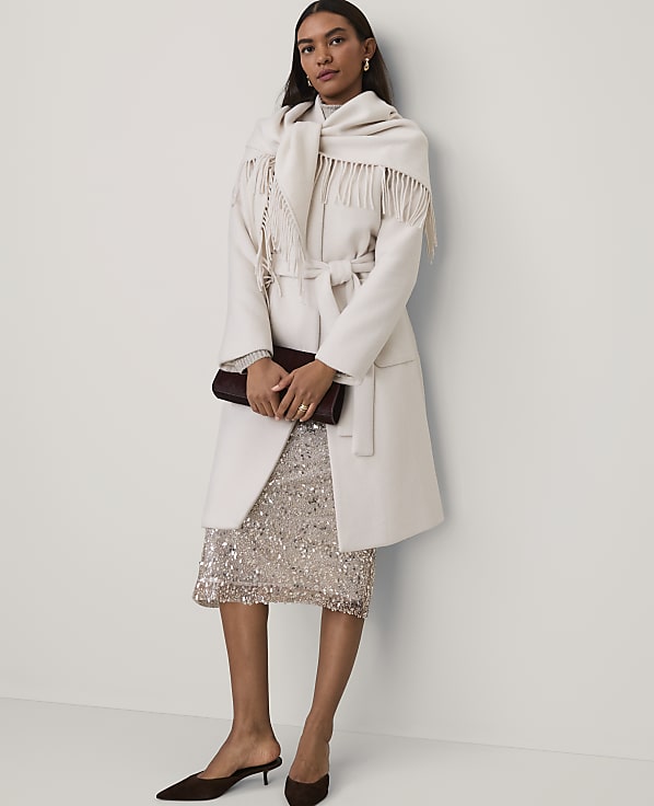 A woman wearing a cream coat, fringed scarf, and sequined skirt stands against a plain white wall.