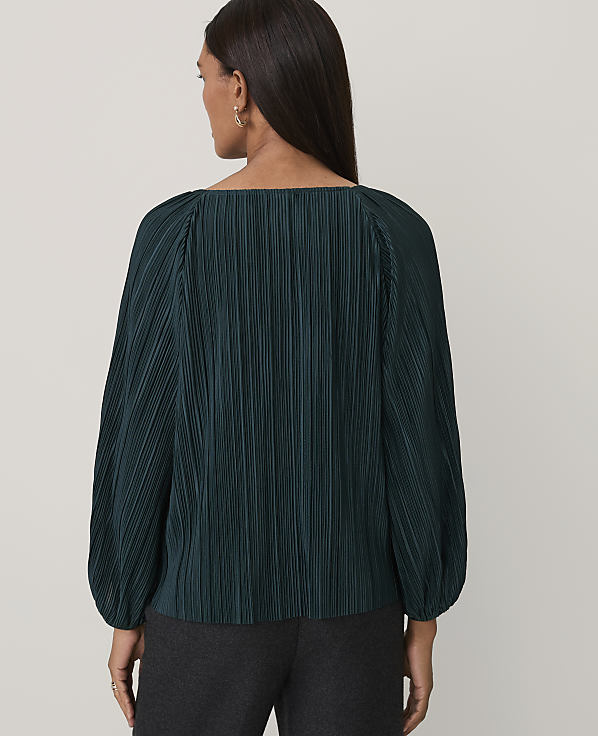 A woman wearing a dark green pleated blouse and charcoal trousers stands against a plain light gray background.