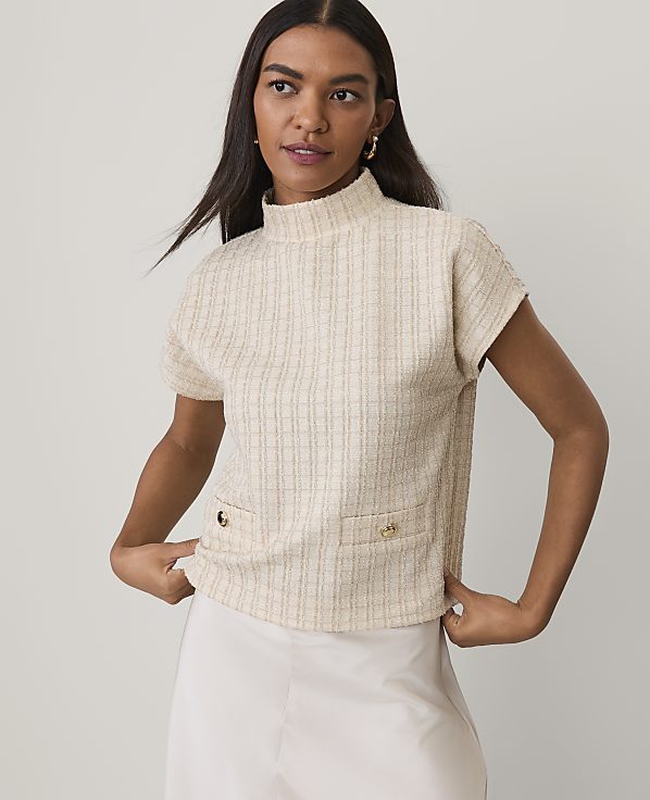 A woman wearing a cream-colored textured short-sleeve top and a white skirt stands against a plain light gray background.