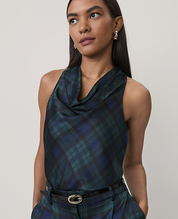 A woman wearing a dark green and blue plaid cowl-neck top and matching trousers stands against a plain, light-colored background.