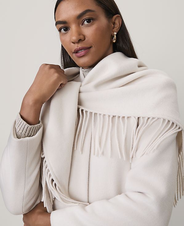 A woman wearing a white coat and a fringed white scarf poses against a plain, light-colored background.
