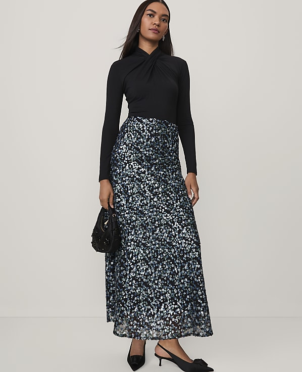 A woman wearing a black long-sleeved top and a shimmering blue and silver sequined skirt stands against a plain, light-colored background.