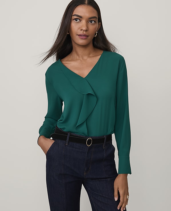 A woman wearing a green ruffle-front blouse and dark blue jeans stands against a plain light gray background.