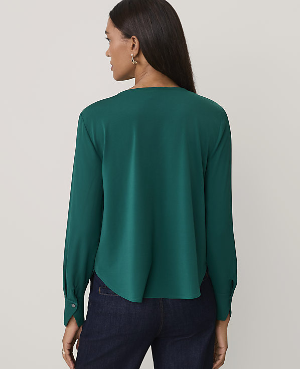 A woman wearing a dark green long-sleeved blouse and navy blue jeans stands against a plain, light-colored background.
