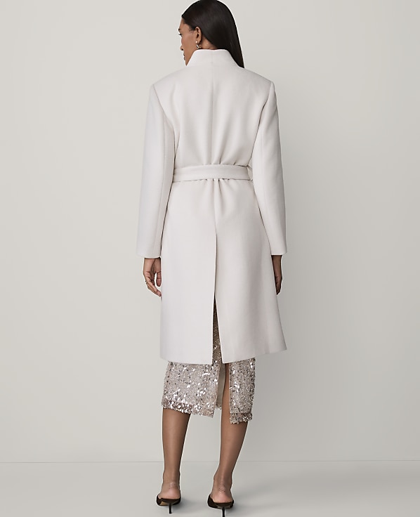 A woman stands with her back to the camera wearing a long white belted coat and a sequined skirt against a plain light grey background.