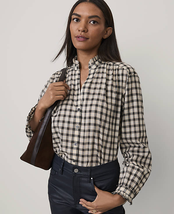 A woman wearing a plaid shirt and dark pants stands against a plain, light-colored background.