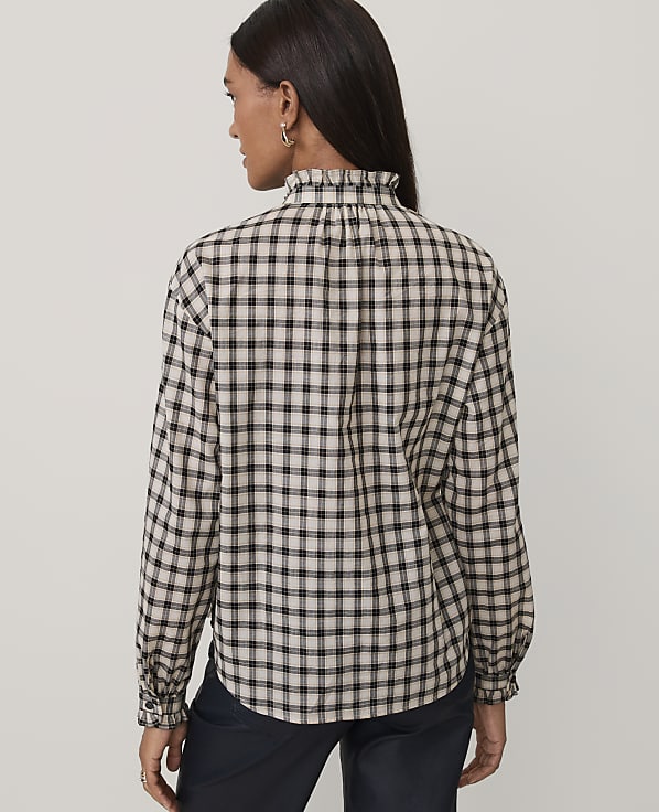 A woman wearing a black and white checkered shirt and dark trousers stands against a plain light gray background.