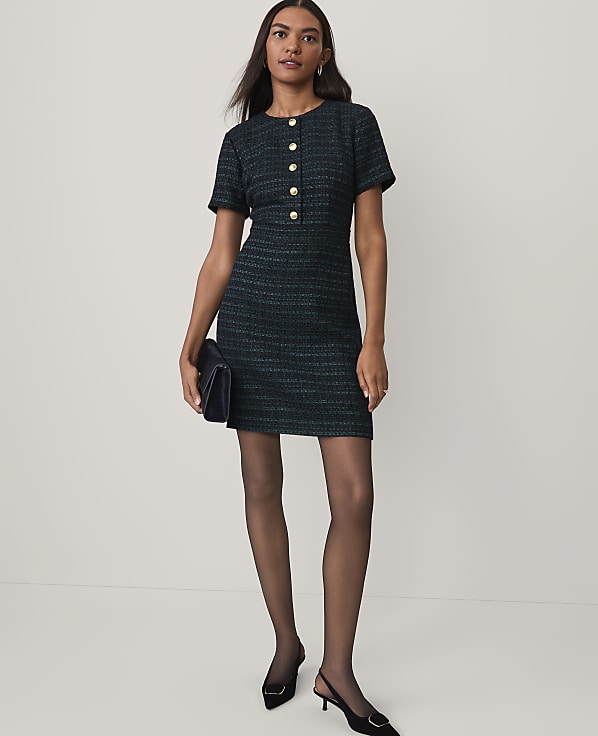 A woman wearing a dark green tweed dress and black heels stands against a plain white background.