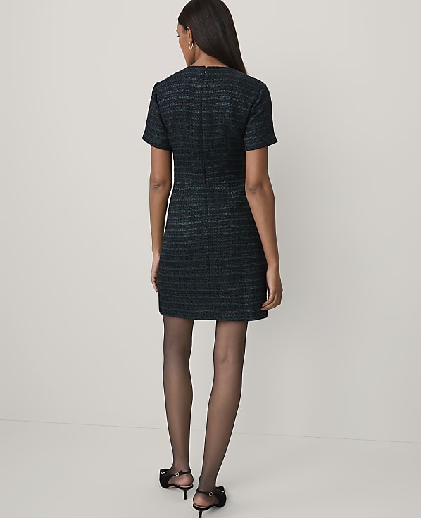 A woman stands with her back to the camera wearing a short-sleeved, textured dark blue dress and black heels against a plain white background.