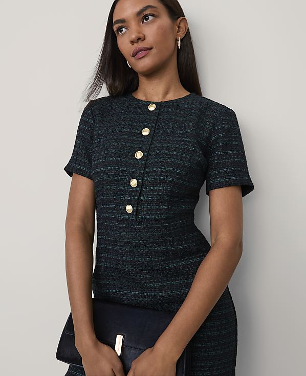 A woman wearing a dark teal textured dress with gold buttons holds a navy clutch against a plain light gray background.