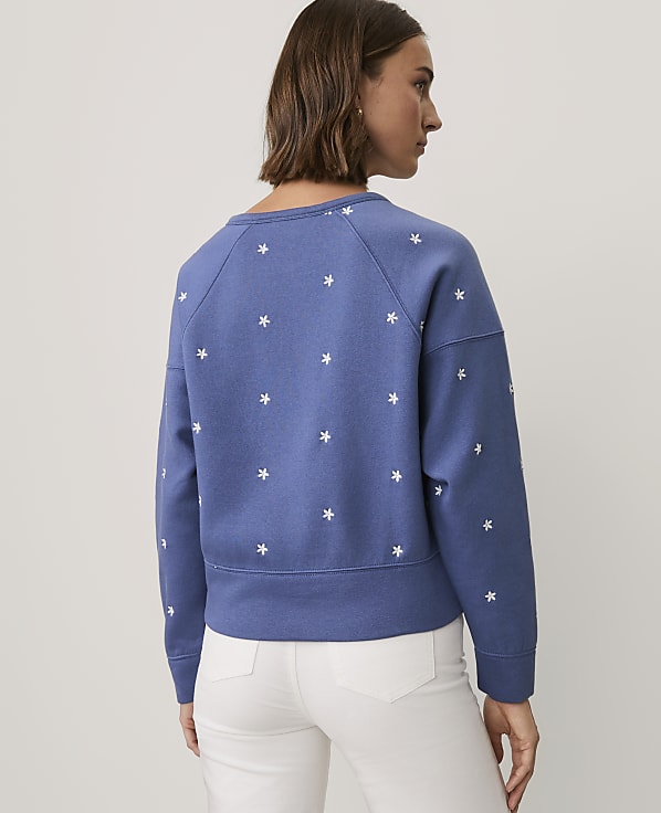 A woman wearing a blue floral-patterned sweatshirt and white pants stands against a plain light gray background.