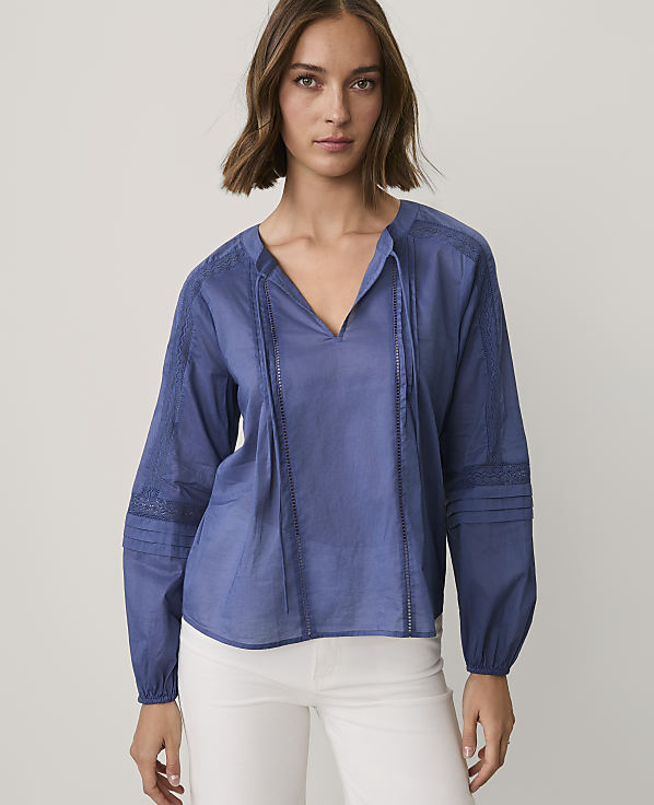 A woman wearing a blue long-sleeved blouse and white pants stands against a plain, light-colored background.