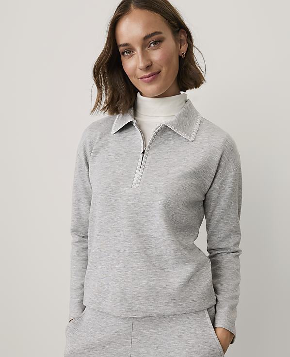 A woman wearing a grey quarter-zip pullover and matching pants stands against a plain, light-colored background.