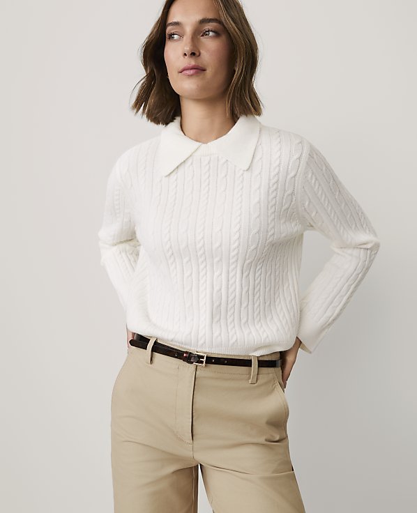 A woman wearing a white cable-knit collared sweater and khaki pants stands against a plain light gray background.