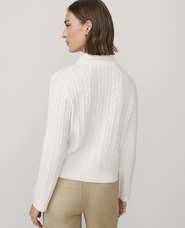 A woman wearing a white cable-knit sweater and khaki pants stands against a plain, light gray background.