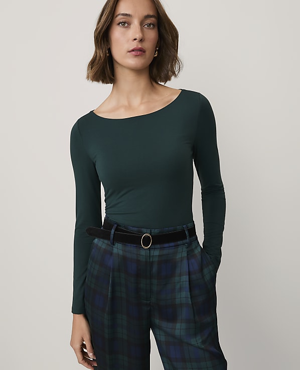 A woman wearing a dark green long-sleeved top and plaid trousers stands against a plain, light-colored background.