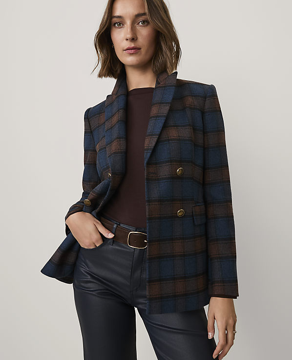 A woman wearing a blue and brown plaid blazer, dark brown top, and black leather pants stands against a plain light-colored background.