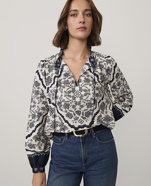A woman wearing a patterned white and navy blouse and blue jeans stands against a plain light gray background.