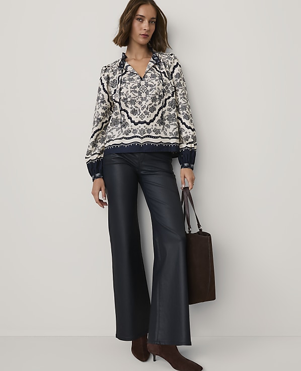 A woman wearing a patterned blouse and navy leather trousers stands against a plain white background while holding a brown tote bag.