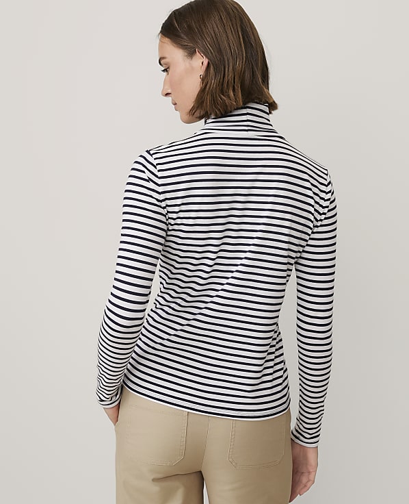 A woman wearing a black and white striped long-sleeved shirt and khaki pants stands against a plain light gray background.