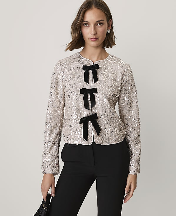 A woman wearing a sequined champagne-colored jacket with black bow accents and black trousers stands against a plain light gray background.