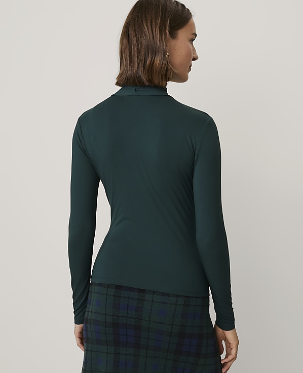 A woman wearing a dark green long-sleeved top and a matching plaid skirt stands against a plain, light-colored background.