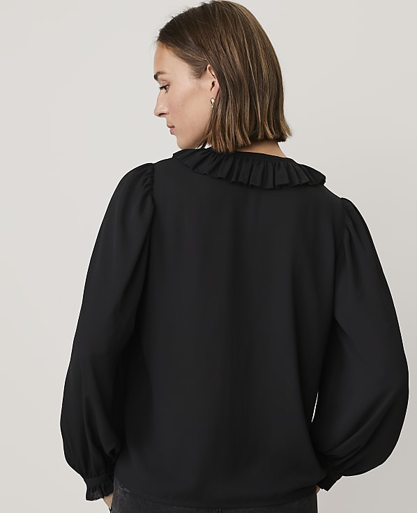 A woman wearing a black ruffled blouse stands with her back to the camera against a plain light gray background.
