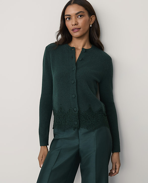 A woman wearing a dark green cardigan with lace detailing and matching trousers stands against a plain, light-colored wall.