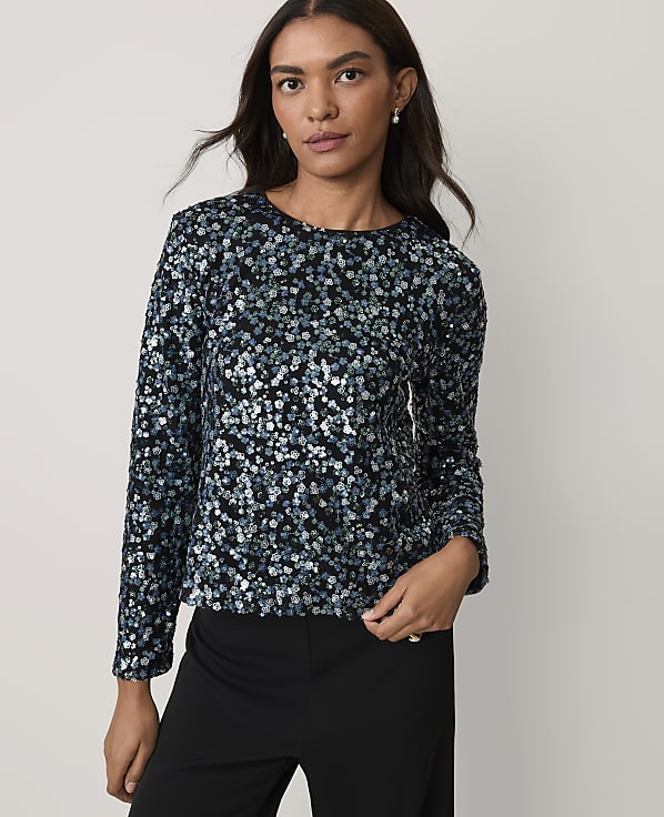 A woman wearing a shimmering blue and black sequined top and black trousers stands against a plain, light-colored background.
