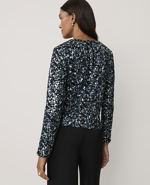 A woman wearing a shimmering blue and black sequined top and black trousers stands against a plain, light-colored background.