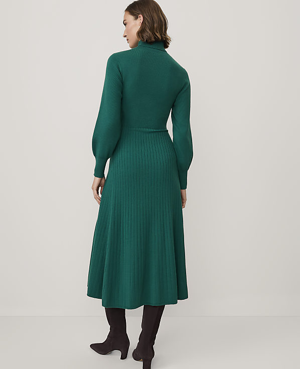 A woman wearing a long-sleeved green ribbed knit dress and dark brown boots stands against a plain white background.