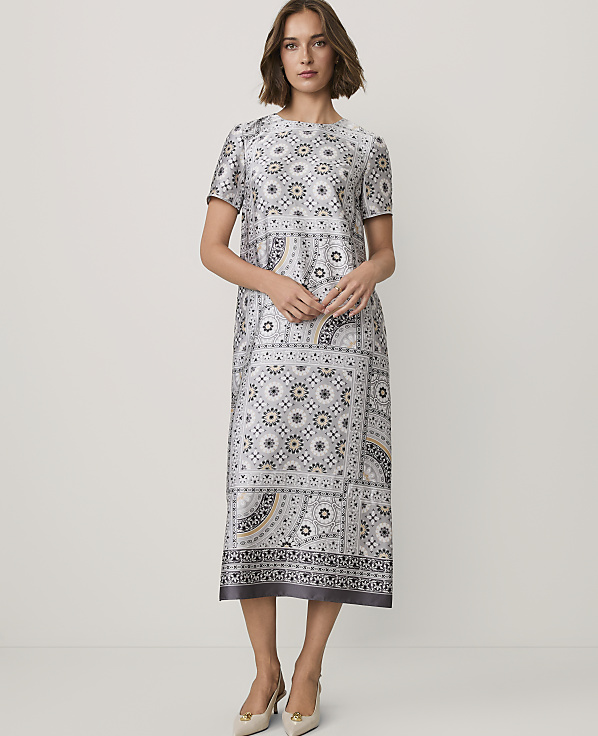 A woman wearing a patterned grey midi dress and beige heels stands against a plain white background.
