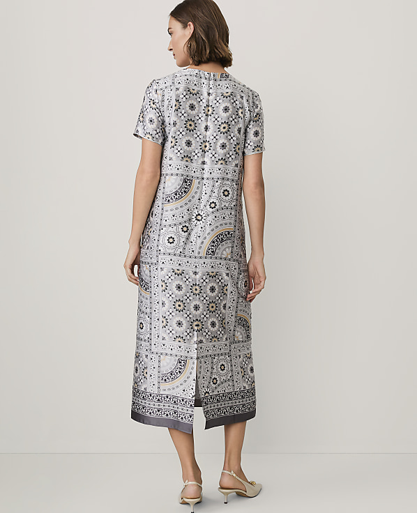 A woman stands with her back to the camera wearing a patterned grey midi dress and beige heels against a plain white background.