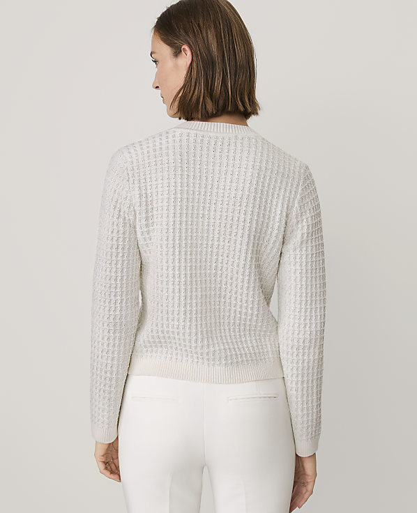 A woman wearing a white textured sweater and white pants stands against a plain light gray background.
