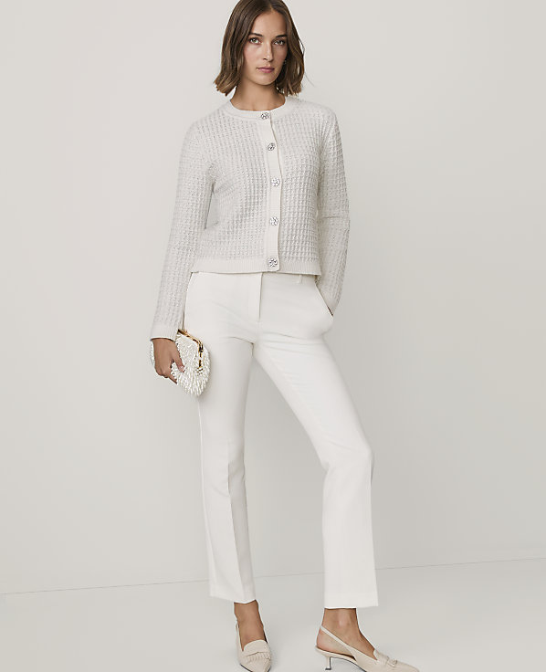 A woman wearing a white textured cardigan, white trousers, and beige heels stands against a plain white background.