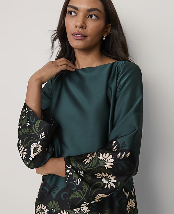 A woman wearing a dark green satin dress with floral patterned sleeves poses against a plain light grey background.