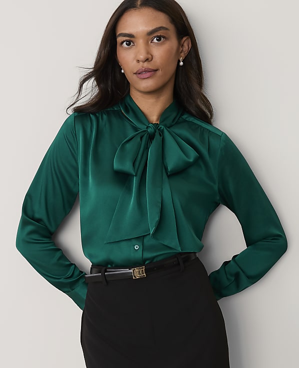 A woman wearing a green satin pussy-bow blouse and black trousers stands against a plain light-grey background.