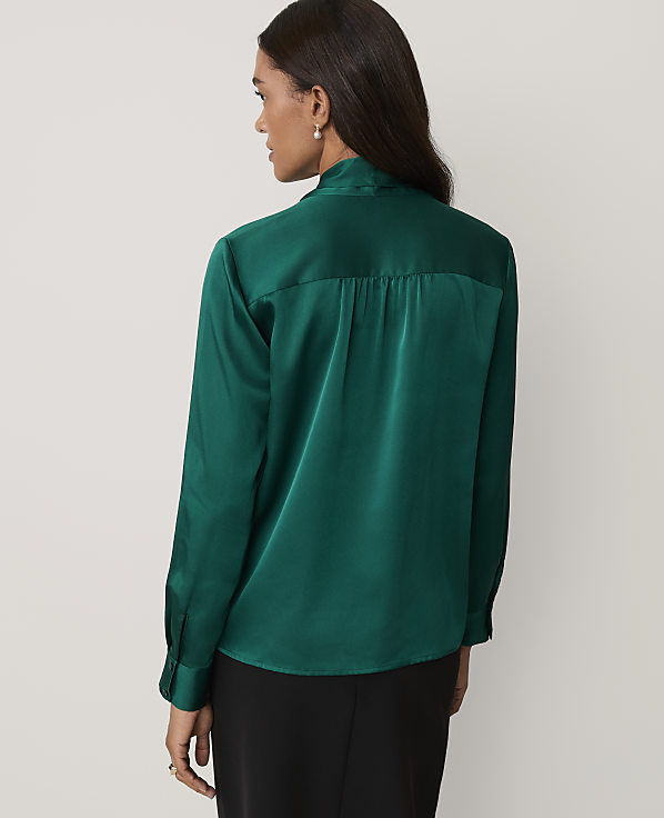 A woman wearing a green satin shirt and black trousers stands against a plain light gray background.