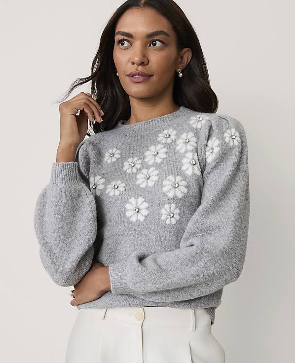 A woman wearing a grey floral-patterned sweater and white trousers poses against a plain light-colored background.