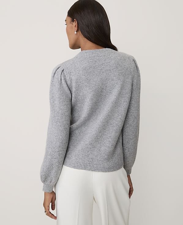 A woman wearing a grey puff-sleeved sweater and white trousers stands against a plain, light-colored background.